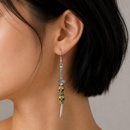 A close-up side profile of a woman with short black hair wearing a dangling earring. The earring features a silver-toned dagger motif with a gold-toned skull accent and a white curved blade tip, set against a neutral gray background.