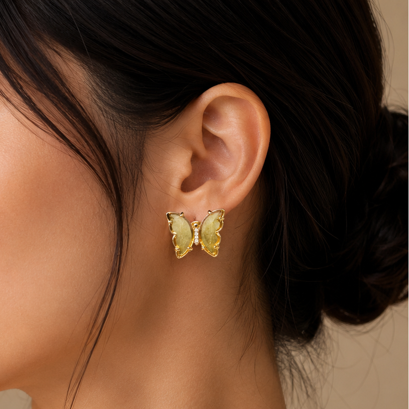 A close-up side profile of a woman wearing a small yellow butterfly-shaped stud earring with gold-toned metal borders, against a soft beige background.