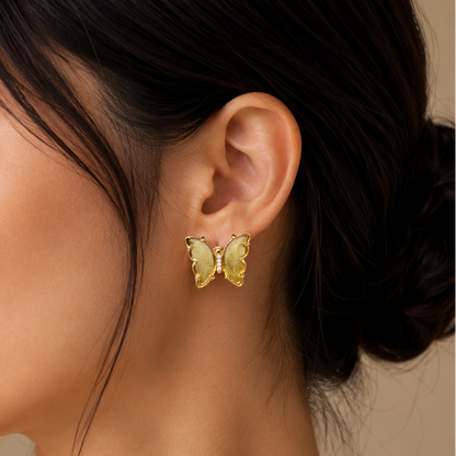 A close-up side profile of a woman wearing a small yellow butterfly-shaped stud earring with gold-toned metal borders, against a soft beige background.