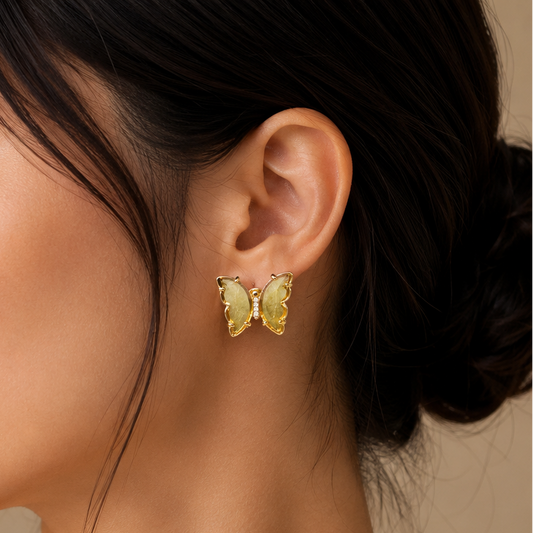 A close-up side profile of a woman wearing a small yellow butterfly-shaped stud earring with gold-toned metal borders, against a soft beige background.