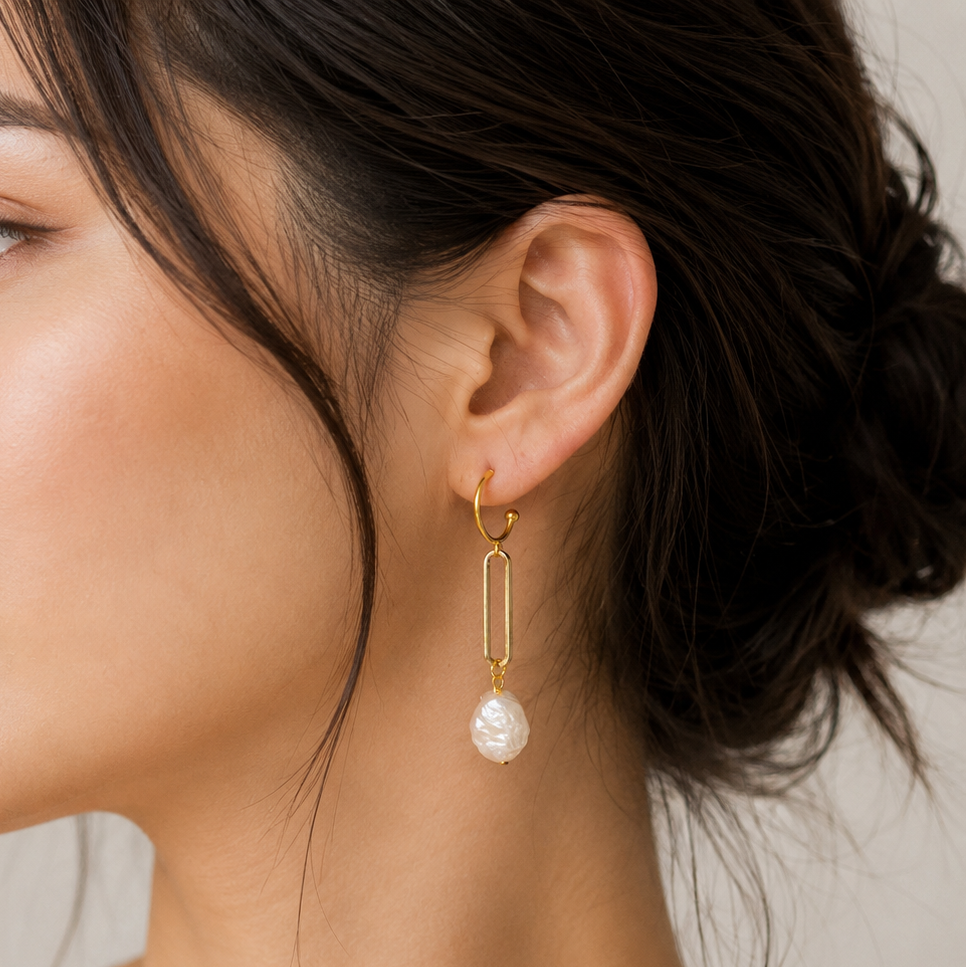 A close-up side profile of a woman wearing a delicate gold huggie hoop earring with a dangling bar and a single white freshwater pearl drop, set against a soft neutral background.