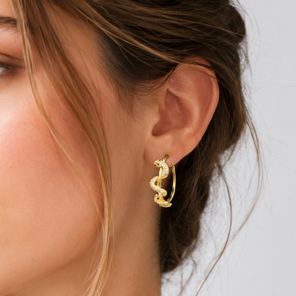 A close-up of a woman wearing a small gold hoop earring with a snake design, paired with a delicate gold chain necklace and a white top against a light gray background.