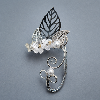 A silver-toned wire art piece featuring a leaf, flowers, and pearls, displayed on a textured gray background.