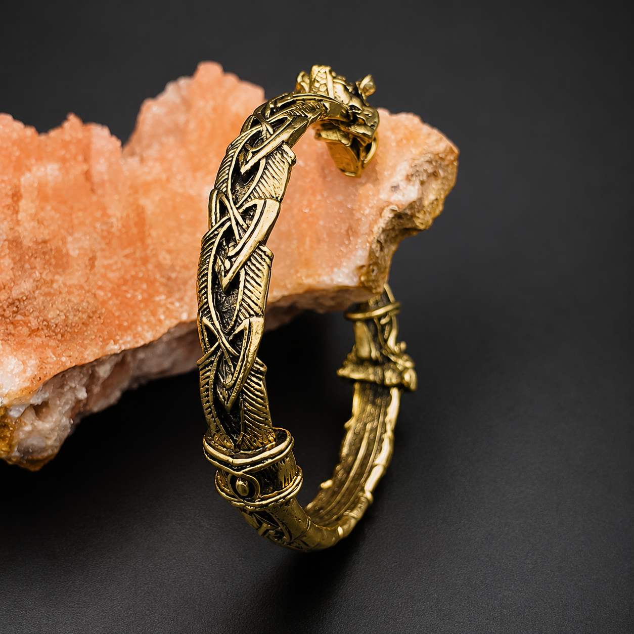 An ornate gold-toned Celtic knot bangle bracelet with a dragon head clasp, displayed on a textured orange rock against a dark gray background.