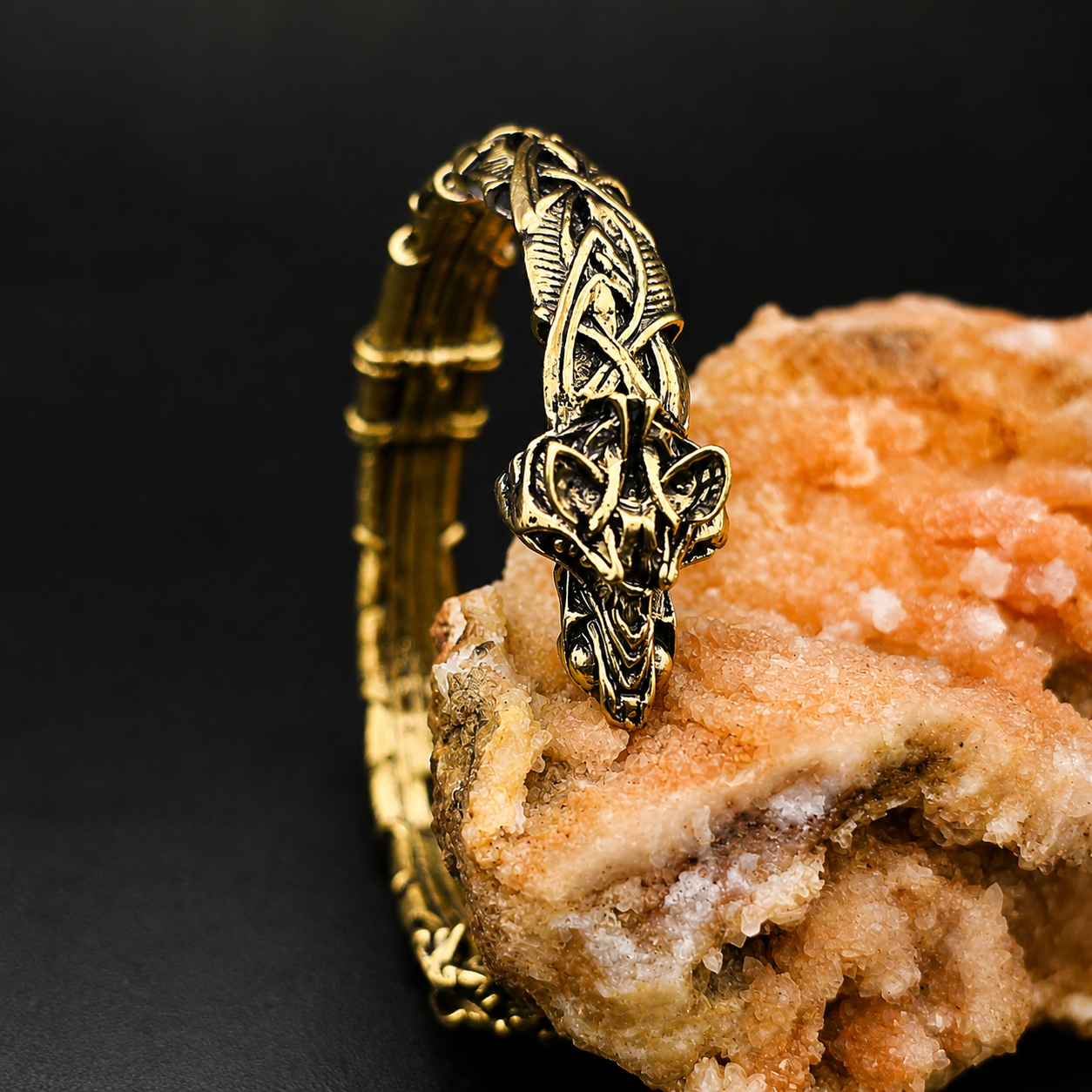 An ornate gold-toned metal cuff bracelet with a detailed Celtic knot and wolf head design, displayed on a textured orange rock against a dark background.