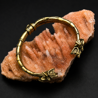 A gold-toned metal bangle bracelet with a textured, segmented design and butterfly charms, displayed on a pinkish-orange crystal cluster against a black background.