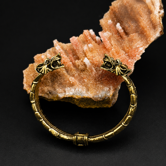 An antique-style gold bangle bracelet with decorative bow details on the clasps, displayed on a textured orange rock against a black background.