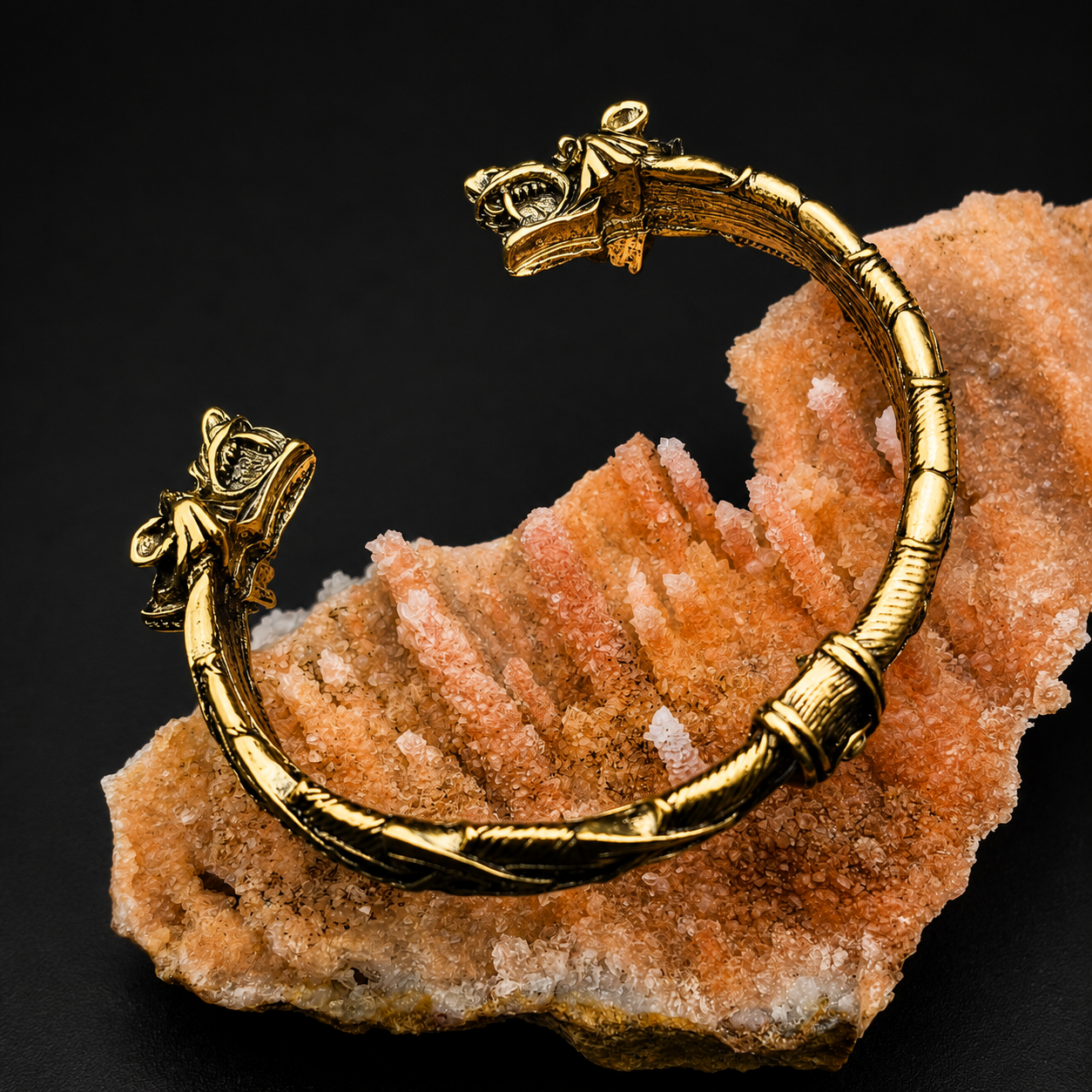 A gold-toned open cuff bracelet with sculpted animal head ends, displayed on a peach-colored crystal cluster against a black background.