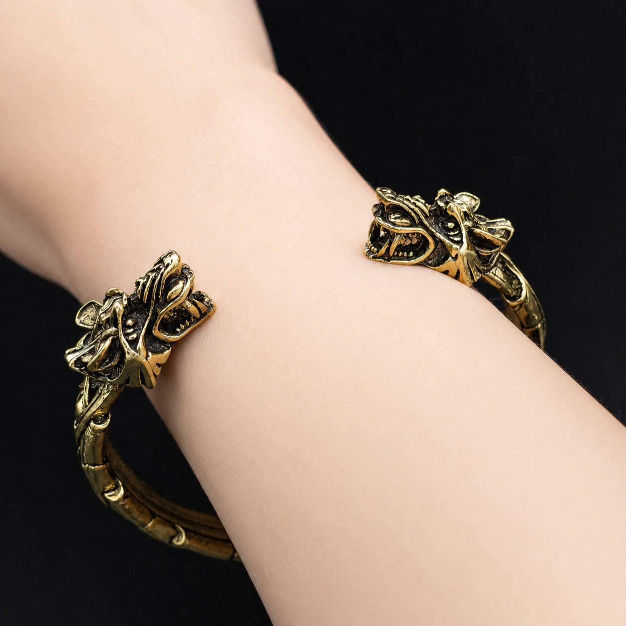 A close-up of a person's wrist wearing a gold-toned metal cuff bracelet with ornate dragon head details on both ends, set against a solid black background.