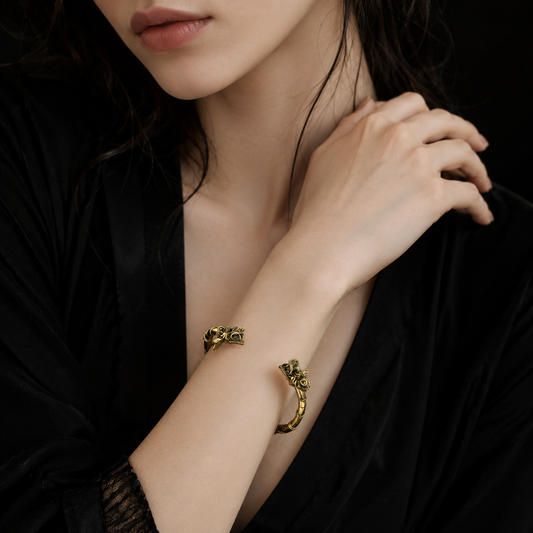 A close-up of a model wearing two gold-toned textured bracelets on her wrist, dressed in a black satin robe against a dark background.