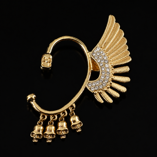 Gold-toned C-shaped hoop earring with a wing design, skull detail, and dangling bell charms, set against a solid black background.