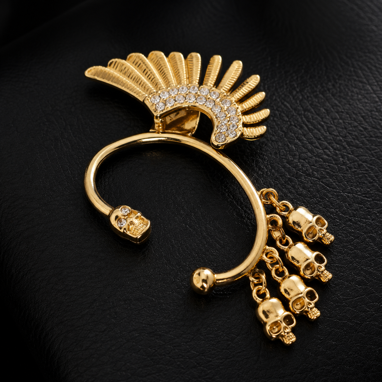 Gold-toned gothic hoop earring featuring a feathered headdress design, skull details, and dangling skull charms, embellished with small clear rhinestones, set against a black textured background.