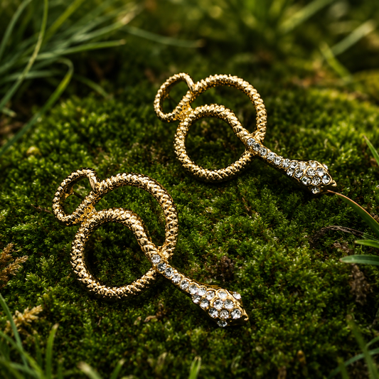 A pair of gold snake-shaped dangle earrings with clear rhinestone accents, displayed on a bed of green moss outdoors.