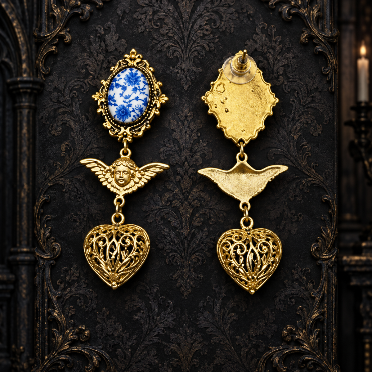 A pair of ornate gold-toned gothic earrings displayed against a dark patterned backdrop. One earring features a blue and white oval stone in a gold frame, while the other shows the back of the piece with a gold wing and heart dangle.