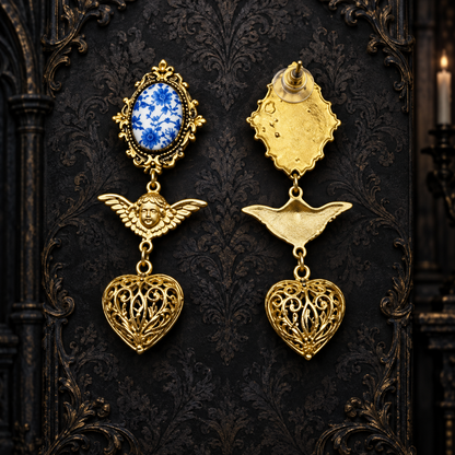 A pair of ornate gold-toned gothic earrings displayed against a dark patterned backdrop. One earring features a blue and white oval stone in a gold frame, while the other shows the back of the piece with a gold wing and heart dangle.