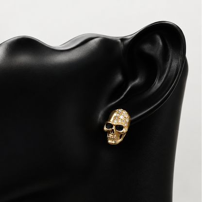 A gold skull stud earring with clear crystals, shown worn on the ear of a matte black female mannequin bust against a light gray background.