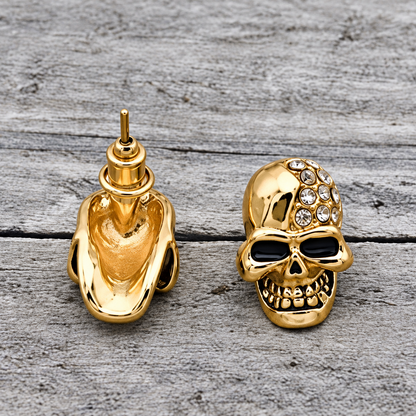 A pair of gold-toned skull stud earrings displayed on a weathered gray wood surface. One earring is shown from the front, featuring blackened eye sockets, a toothy grin, and a textured top adorned with clear rhinestones.