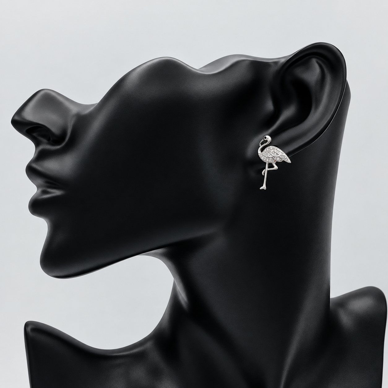 A silver-toned flamingo stud earring displayed on the ear of a matte black female mannequin head against a light gray background.