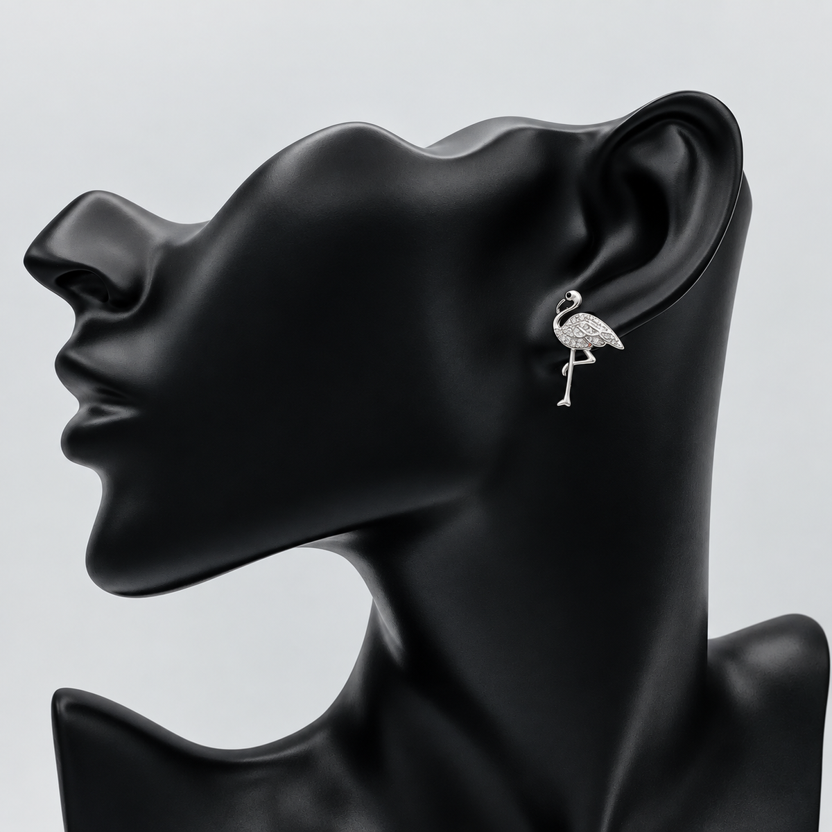 A silver-toned flamingo stud earring displayed on the ear of a matte black female mannequin head against a light gray background.