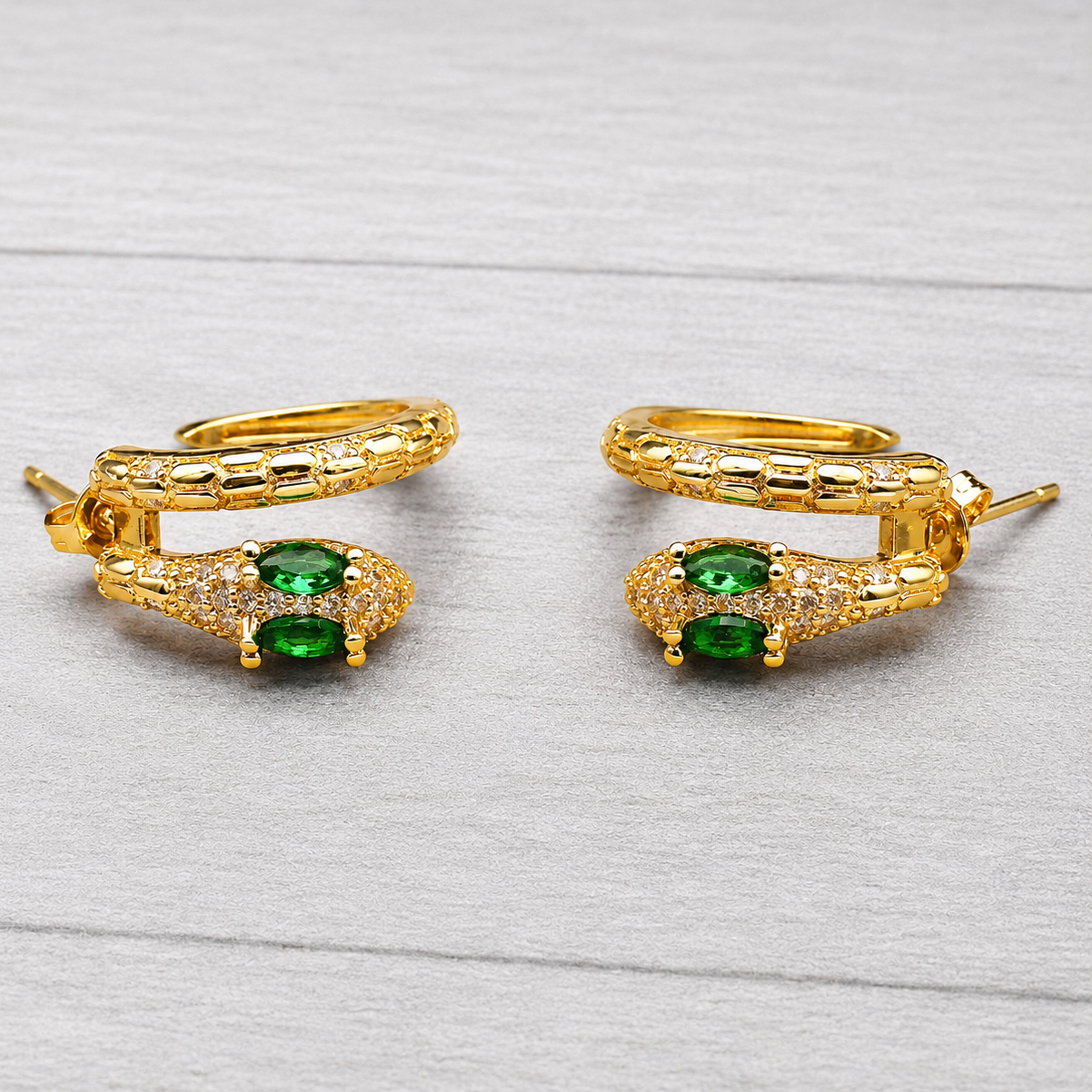 A pair of gold hoop earrings with a snake-head design, featuring green oval gemstones and small clear pavé accents, displayed on a light gray wood-textured background.
