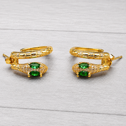 A pair of gold hoop earrings with a snake-head design, featuring green oval gemstones and small clear pavé accents, displayed on a light gray wood-textured background.