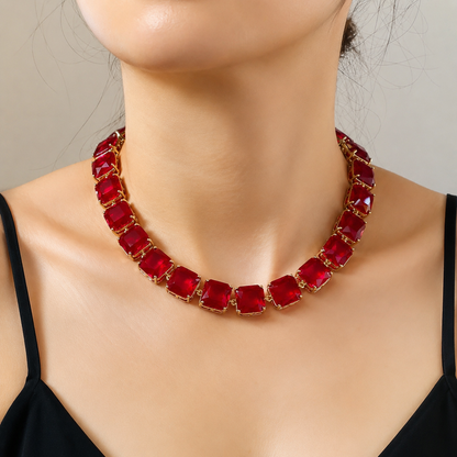 A close-up of a model's neck and upper chest wearing a chunky gold-tone necklace with large, faceted deep red gemstones.