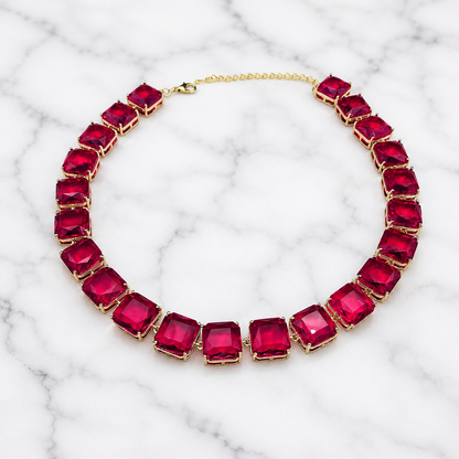 A gold-plated collar necklace featuring large, square-cut deep pinkish-red gemstones, displayed on a white marble background.