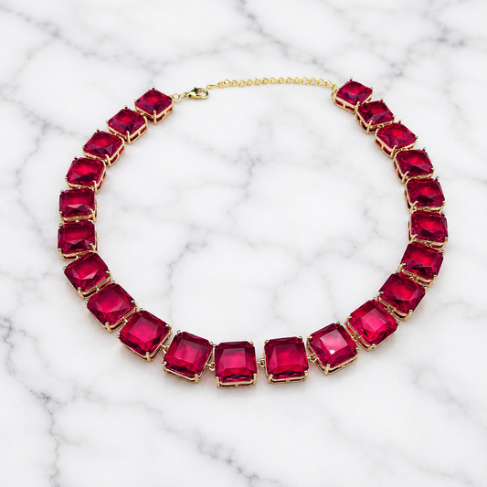 A gold-plated collar necklace featuring large, square-cut deep pinkish-red gemstones, displayed on a white marble background.