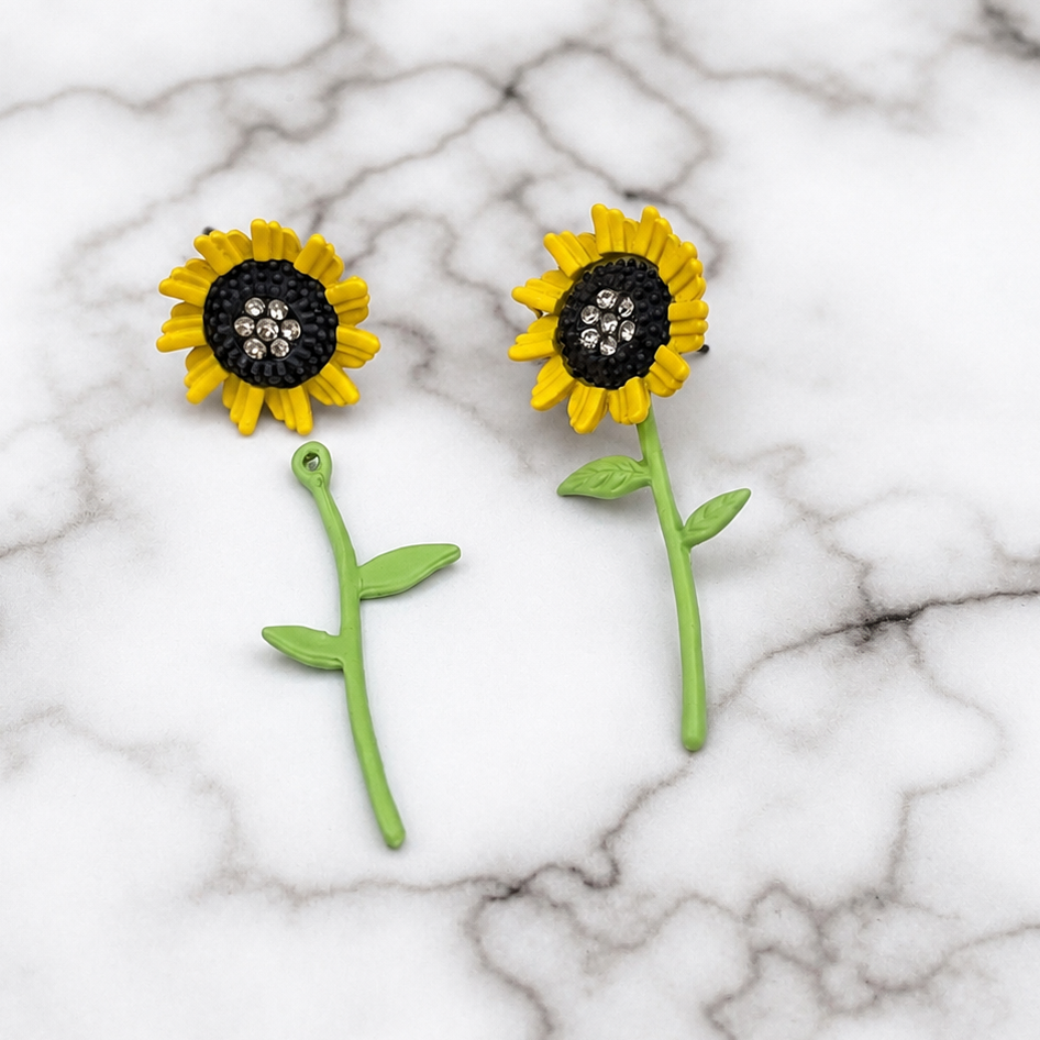 A pair of yellow sunflower stud earrings with black centers and green stems, displayed on a white and gray marble surface.