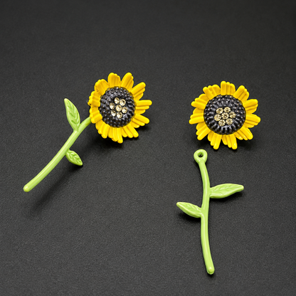 A pair of yellow sunflower stud earrings with black centers and rhinestone accents, shown alongside a matching green stem and leaf charm on a dark gray background.