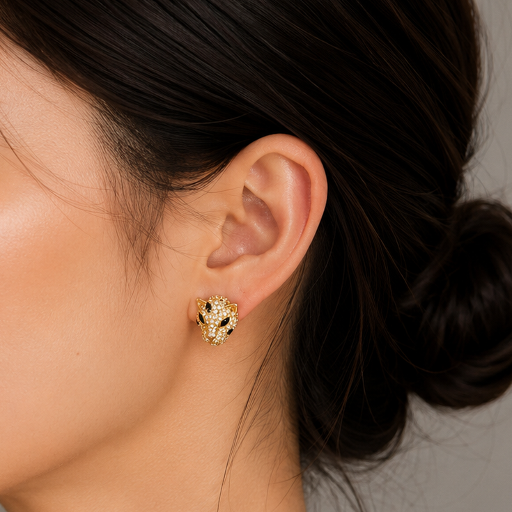 A close-up side profile of a woman with dark hair in a bun, wearing a small textured gold stud earring against a neutral gray background.
