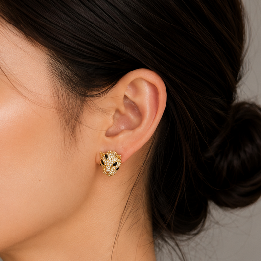 A close-up side profile of a woman with dark hair in a bun, wearing a small textured gold stud earring against a neutral gray background.