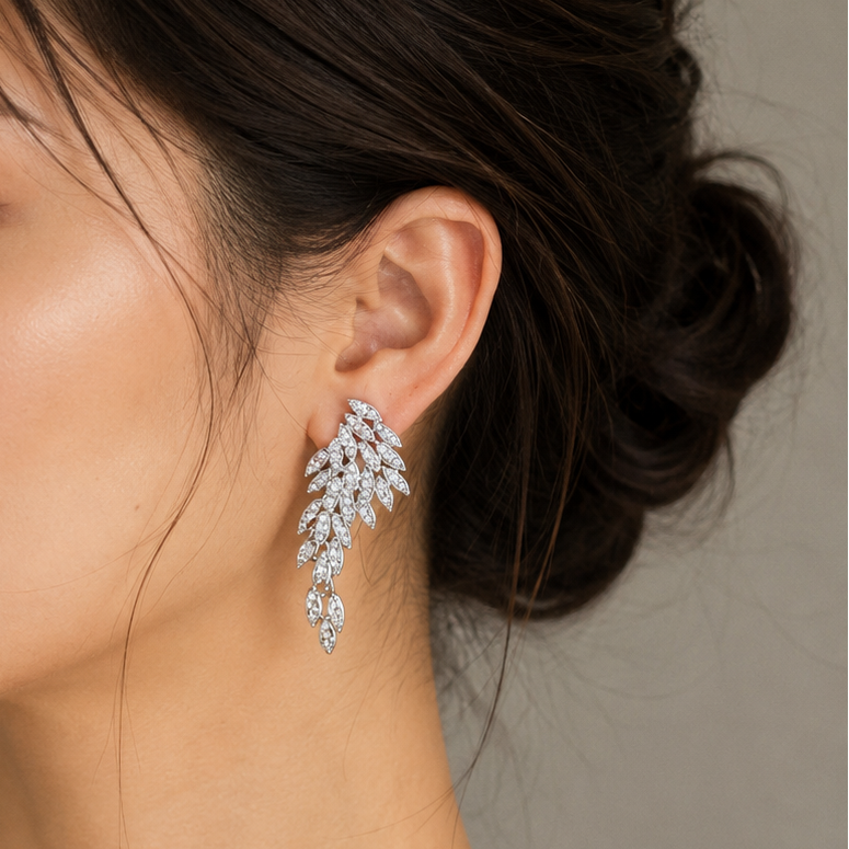 A woman in profile wearing a delicate silver-toned leaf-shaped drop earring set with clear stones, shown against a soft gray background.