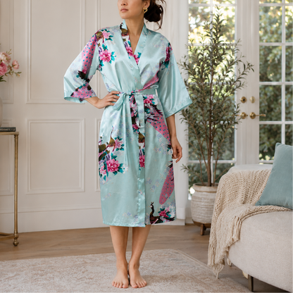 A woman stands barefoot in a bright living room wearing a light aqua satin robe with a pink, purple, and brown floral print. The robe has wide sleeves and a self-tie belt.