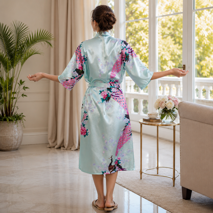 A woman stands with her back to the camera wearing a light blue satin robe with a pink and green peacock print, in a bright, modern living room.