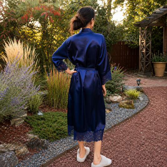 A woman wearing a shiny navy blue satin robe with lace trim and white slippers, standing in a garden backyard setting.