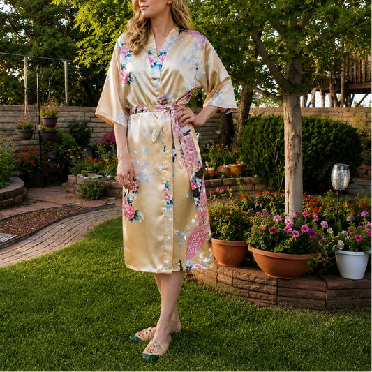 A woman stands on a grassy backyard garden lawn wearing a beige satin kimono robe with colorful floral prints and a matching tie belt.