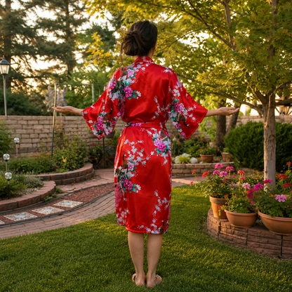 A woman stands barefoot in a backyard garden, viewed from behind, wearing a bright red kimono-style robe with a large pink, purple, and white floral print. She has her arms outstretched, with trees, grass, and potted flowers in the background.
