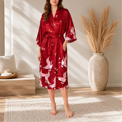 A woman stands barefoot in a sunlit room wearing a red satin-like robe with white crane prints and small floral motifs. The robe has wide sleeves, a V-neck, and a self-tie belt.