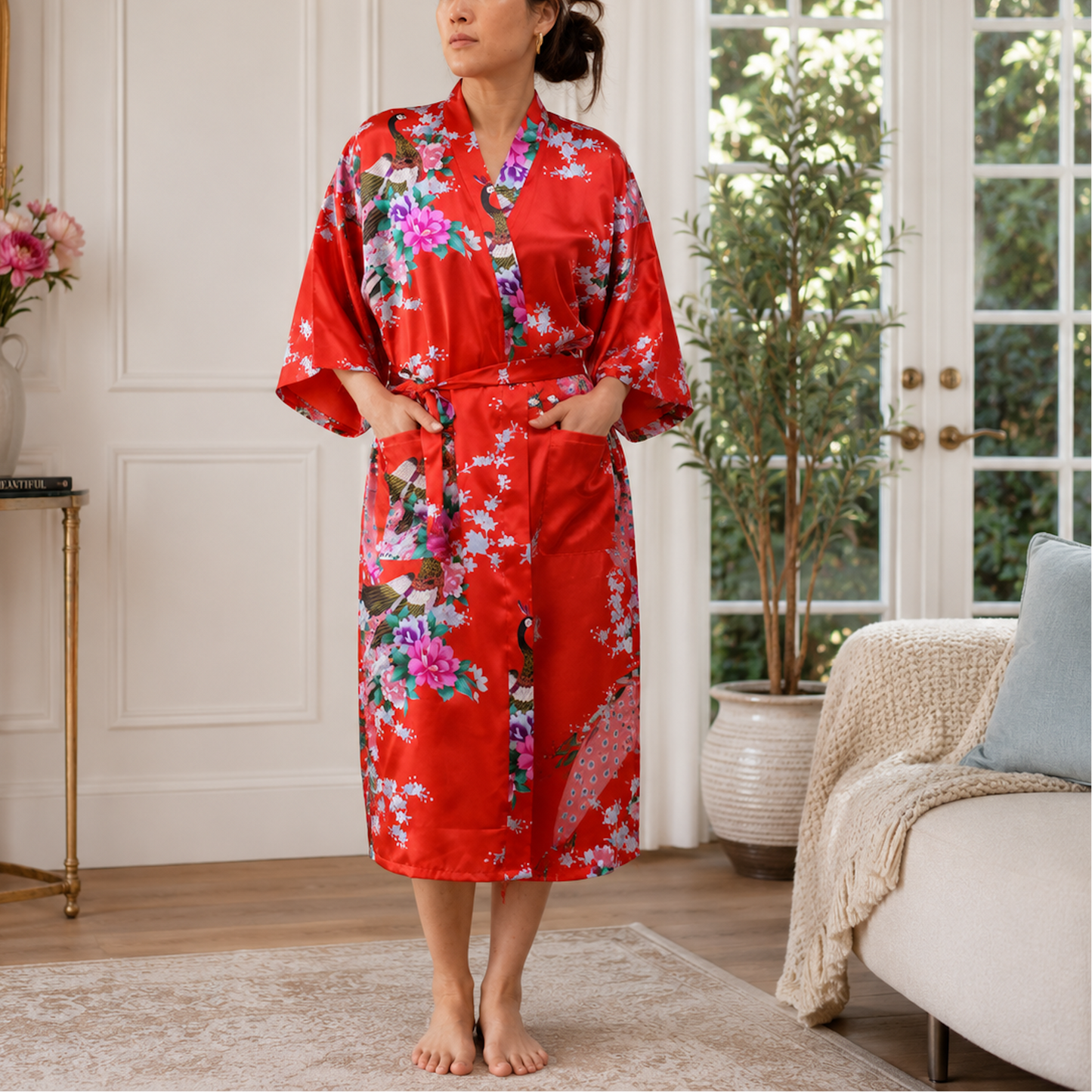 A woman stands barefoot in a bright, elegant room wearing a red satin kimono-style robe with a colorful floral print and a matching tie belt.