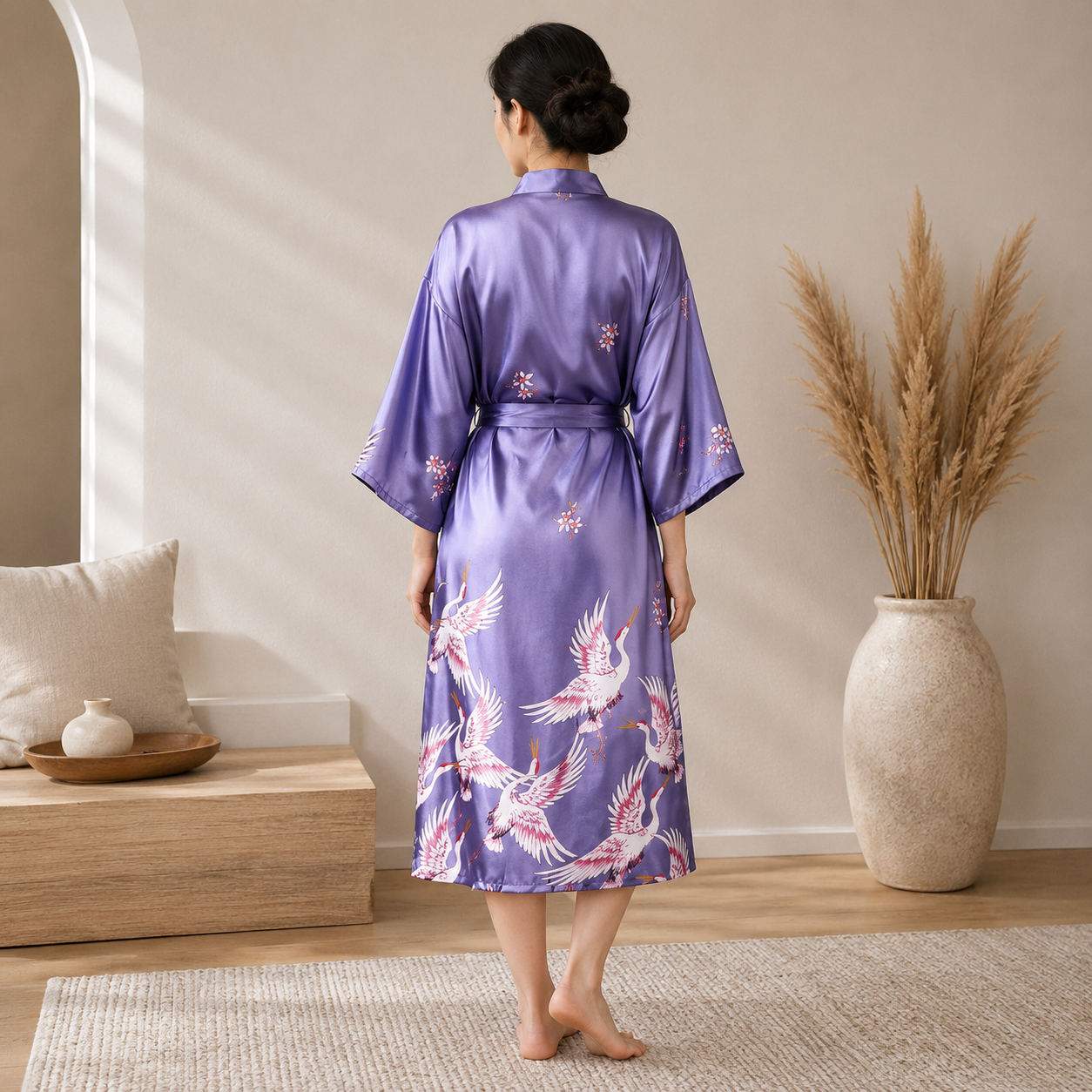 A woman stands with her back to the camera wearing a lavender-purple satin kimono robe with pink and white crane prints. The setting is a beautifully styled room with a wooden bench, decorative pillows, a large vase of dried pampas grass, and a light-colored rug.