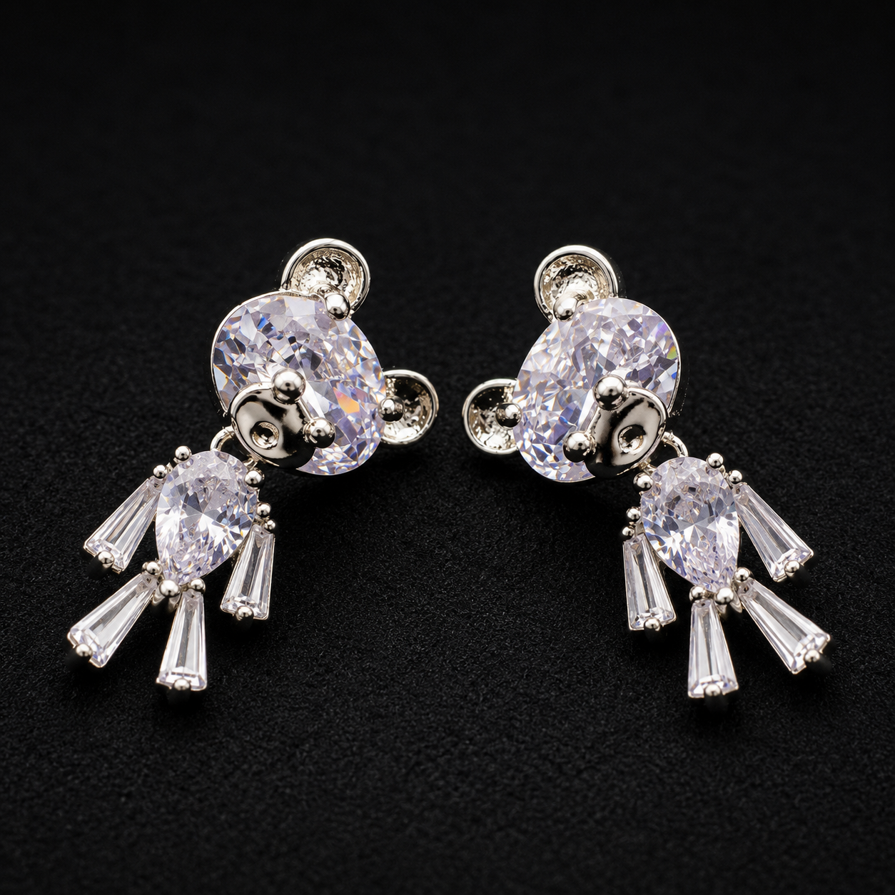 A pair of silver-tone dangle earrings featuring large oval clear crystals on the top and teardrop-shaped clear crystals dangling below, displayed on a textured black background.