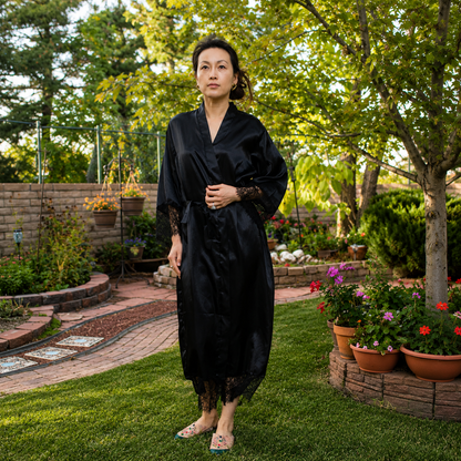 A woman wearing a black satin-like robe with lace sleeves and hem, standing in a lush green backyard garden.