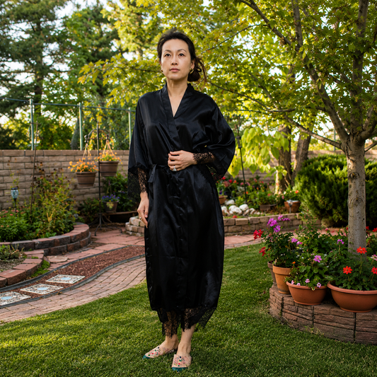 A woman wearing a black satin-like robe with lace sleeves and hem, standing in a lush green backyard garden.