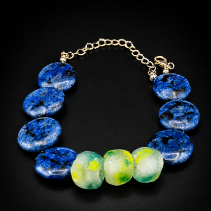 A beaded bracelet featuring seven  blue jasper stones and three translucent green and yellow agate beads, suspended on a gold-tone chain with a lobster clasp, set against a solid black background.