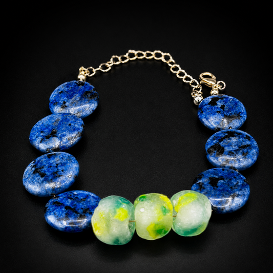A beaded bracelet featuring seven  blue jasper stones and three translucent green and yellow agate beads, suspended on a gold-tone chain with a lobster clasp, set against a solid black background.