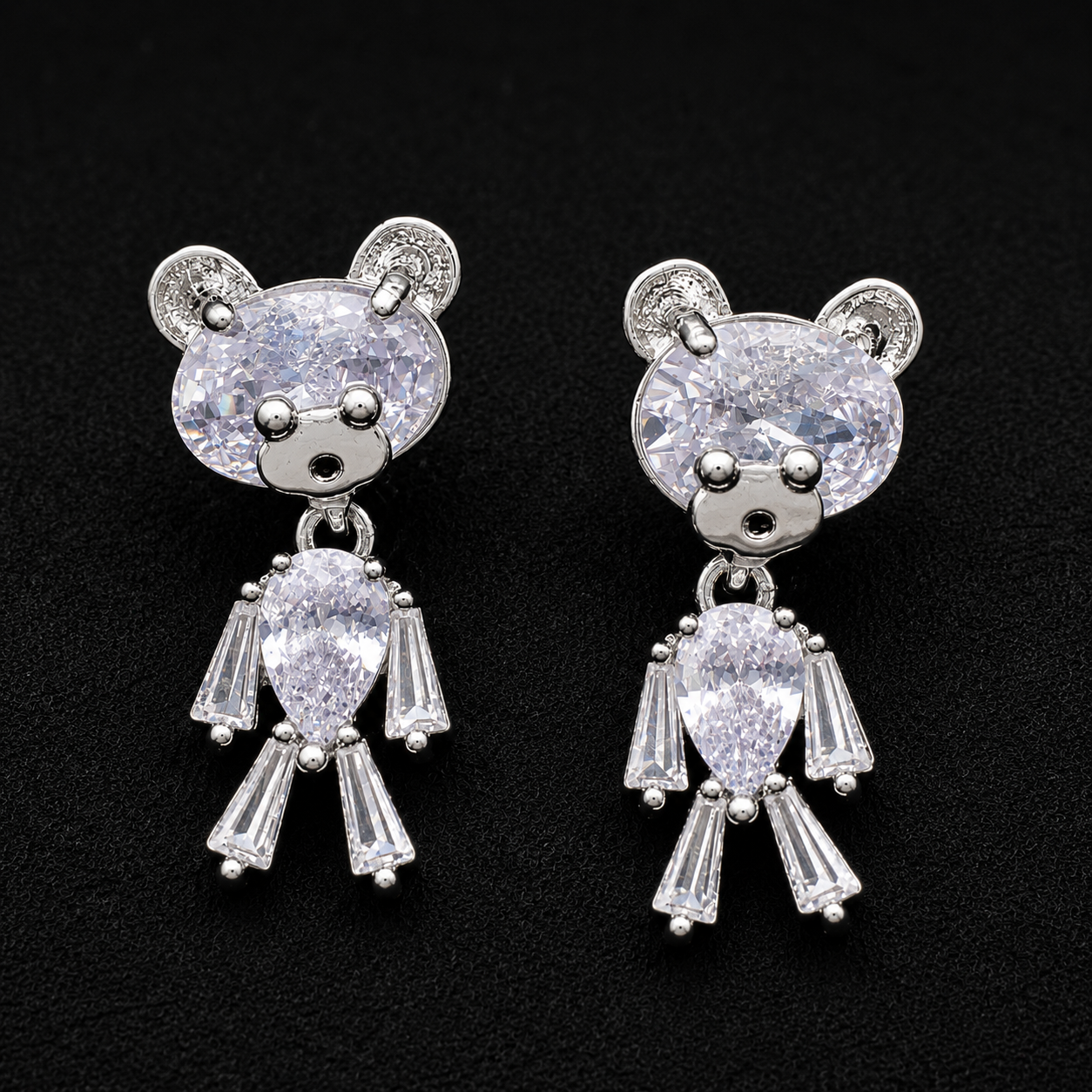 A pair of silver-toned teddy bear stud earrings with clear faceted gemstones, displayed on a textured black background.