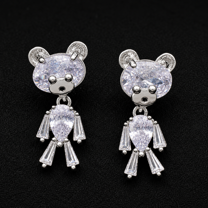 A pair of silver-toned teddy bear stud earrings with clear faceted gemstones, displayed on a textured black background.