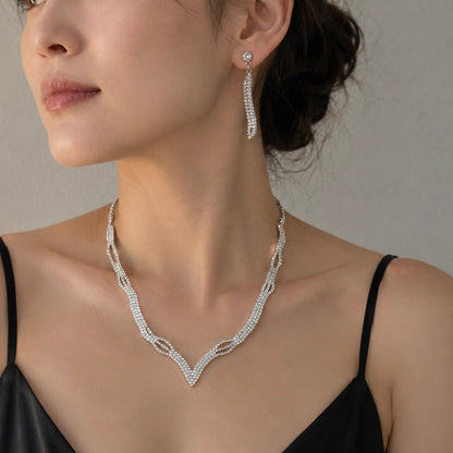 A close-up of a woman wearing a silver V-shaped necklace and matching drop earring, both adorned with sparkling clear stones, against a neutral gray background.