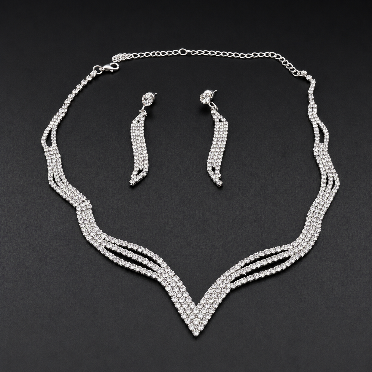 A silver-tone jewelry set featuring a multi-strand V-shaped necklace and matching drop earrings, both adorned with clear rhinestones, displayed on a dark background.